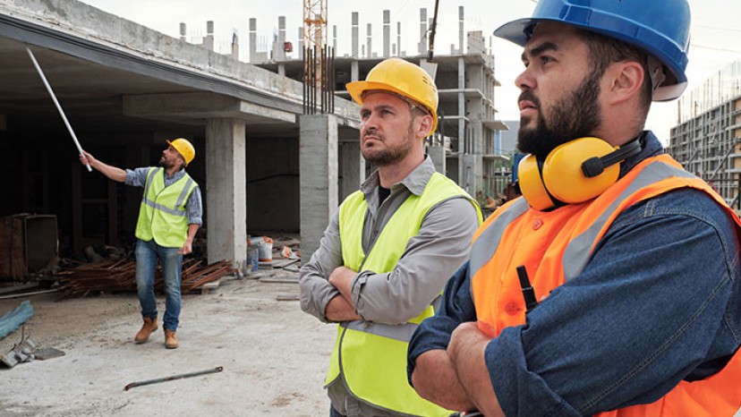 Zwei Kollegen mit Schutzhelmen und -westen stehen auf einer Baustelle, während im Hintergrund ein weiterer Mann steht.